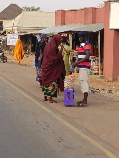 The image captures a vibrant street scene, likely in a town or village, with several individuals and roadside stalls. The primary focus is on two people interacting near a purple wheeled trolley filled with goods.
In the foreground, on the left side of the trolley, stands a woman wearing a dark red hijab that covers her head and shoulders, paired with a long green skirt featuring a gold and black pattern. She is seen with her hands near her face, possibly eating a small item or engaged in conversation. On the right side of the trolley, a man stands, wearing a blue and white striped short-sleeved t-shirt, light-colored shorts that reach his knees, and a blue cap. He is looking towards the woman, and his body language suggests he might be presenting the items on the trolley or engaged in a discussion with her. The purple trolley is a small, compact unit with wheels, and on top of it sits a clear plastic bucket overflowing with numerous small, colorful items, which appear to be candies or small toys, arranged in distinct blocks of color.
Behind the woman in red, another woman is partially visible. She is dressed in a dark blue patterned dress or outfit with a matching head covering. Further down the road on the far left, an individual shrouded entirely in a bright orange garment is seen walking away from the camera.
The background features several single-story buildings that appear to be shops or market stalls, characterized by corrugated metal roofs and painted walls, predominantly in a light red or terracotta color. One stall on the right shows shelves stocked with various bottled drinks. On the far left, a white signboard is visible, partly obscured by the orange-clad figure. The legible text on the signboard reads "ST. PETER'S ANGLICAN CHURCH," with additional smaller text below, possibly detailing church activities or service times. A motorcycle is partially visible parked near the signboard.
The scene takes place outdoors during what appears to be daytime, with soft lighting indicating either morning or late afternoon. The ground consists of both asphalt road with a faded yellow dividing line and dusty, unpaved areas off to the sides. The people are dressed in everyday attire, with the women wearing traditional head coverings and long garments, and the man wearing casual clothing and flip-flops. The overall impression is that of a bustling local street market or roadside vendor interaction.