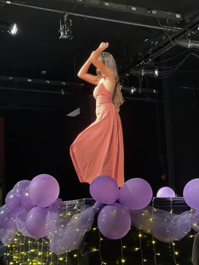 The image depicts a performance setting within an unconfirmed venue in Palos Verdes Estates, United States. A single individual, observed from a side profile, stands center-frame on what appears to be an elevated stage. The individual has long, wavy, light-colored hair extending down their back and is wearing a red and white gingham-patterned maxi dress with a halter neckline and an open back design. Their arms are raised above their head, bent at the elbows, with hands positioned together, suggesting a dynamic pose or dance movement.

In the foreground, a dark structure or railing is decorated with multiple matte purple balloons, purple sheer fabric (tulle), and warm-toned string lights. The balloons partially obstruct the lower portion of the central figure.

The background is dark, revealing a stage environment. Overhead, black structural elements, likely stage rigging, are visible with multiple mounted stage lights or spotlights. White numbers (e.g., "77", "78", "79", "59", "60") are affixed to these structures. The lighting primarily illuminates the central figure and the foreground decorations, with the rest of the background remaining dim.