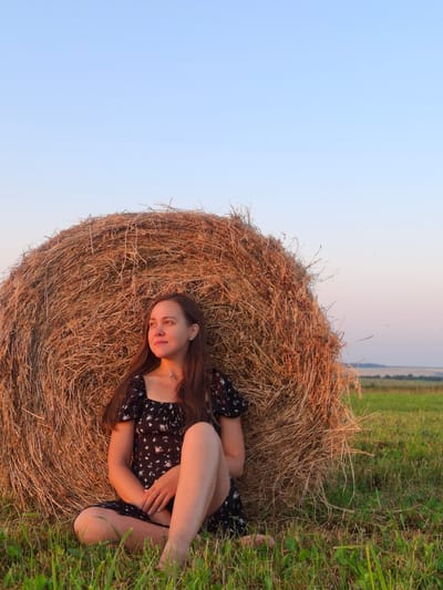 A young woman with long, dark hair sits with her back against a large, round hay bale in a grassy field. She is wearing a short, black dress with a floral pattern. Her legs are crossed, and her hands are resting on her lap. She is looking to her left, with a thoughtful expression on her face. The sky behind her is a clear, pale blue, and the sun appears to be setting, casting a warm glow on the hay bale and the surrounding grass. The background shows a distant horizon line with a hint of trees and a pale, pinkish sky. The scene is located in the city of Perevoznoe, Russia.