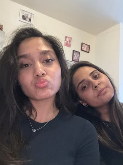 Two women are posing for a selfie in an indoor setting. The woman in the foreground is on the left, with dark hair, and she is puckering her lips. She is wearing a black shirt and a silver necklace. The woman in the background is on the right, also with dark hair. She is looking towards the camera with a neutral expression. Behind the women, there are framed pictures on a cream-colored wall. The overall scene appears to be a casual, personal space, likely a home. There is no discernible indication that this image was taken in Maldegem, Belgium.