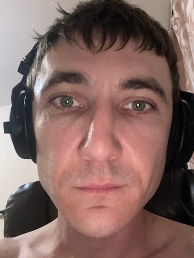 The media file is a close-up, sideways portrait of a person wearing headphones. The subject's face is partially visible, with their eyes and forehead being the most prominent features. They have green eyes and dark hair that appears somewhat disheveled. There is a white mark on their nose, possibly from a product or a smudge. The lighting suggests an indoor setting, and the background is blurry, with hints of fabric and a dark surface, likely furniture. The overall mood is casual and candid, with the subject seemingly looking directly at the camera. No text is visible in the image. The location context provided is Laishevo, Russia.