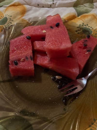 A close-up overhead shot shows a clear plastic plate with several slices of watermelon. The watermelon is bright red with black seeds and some white pith. The plate sits on a cloth with a floral pattern. A silver fork rests on the plate, next to a pile of watermelon seeds and juice. The scene appears to be indoors, possibly on a bed or couch, given the fabric in the background. There is no indication of Garmen, Bulgaria.