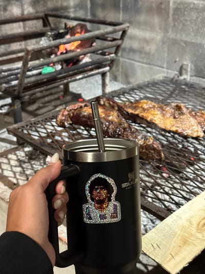 A black Stanley mug with a handle and a sticker is held by a hand with manicured nails. A thin, metallic object protrudes from the mug towards the grilling area. The grill has meat cooking over flames, and a piece of wood rests on the edge. The grill is set on top of gray stone tiles.