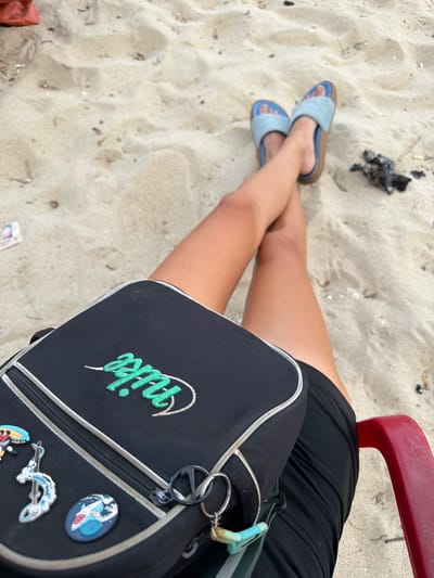 A high-angle shot depicts a person seated on a red chair on a sandy beach in Cam Lâm, Vietnam. The individual is wearing a black outfit and blue sandals. A black handbag with a green "Nike" logo is placed on their lap. The sand is visible in the background, along with some debris.