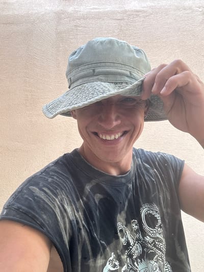 A person wearing a light green bucket hat and a dark t-shirt with a graphic print is looking at the camera and smiling. The hat is tilted, obscuring the person's forehead and eyebrows. The person's right hand is raised, holding the brim of the hat. The t-shirt is dark gray with a distressed, tie-dye pattern and a white graphic of what appears to be an astronaut's helmet and a skeletal hand. The background is a plain, textured, light brown wall. The image is a close-up shot, focusing on the person's face and upper torso. The scene is set outdoors against a building wall.