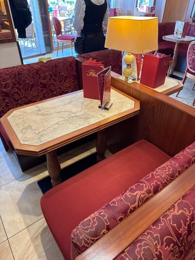 The image shows the interior of a café, likely in Cochem, Germany. A table with a marble top and a wooden frame is partially visible in the foreground. On the table is a menu in a red holder. The café seating includes a red padded booth. A lamp with a yellow shade is present. A waiter wearing a white shirt and black vest is partially visible. In the background, there are other tables, chairs, and patrons. The floor is tiled in light colored. The overall setting suggests a traditional European café environment.