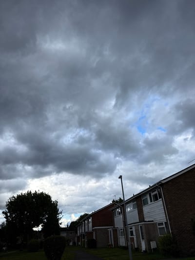 The image depicts a residential street in Hazlemere, United Kingdom, under a dramatic, overcast sky. The foreground features a manicured lawn and a dark silhouette of a large tree on the left. A row of brick terraced houses with white uPVC window frames lines the right side of the frame. A grey lamppost stands prominently in front of the houses. The sky is filled with heavy, dark grey clouds, with a small patch of bright blue sky visible towards the upper right quadrant. The overall impression is one of impending rain or stormy weather.