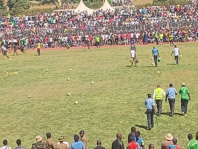 The image depicts an outdoor football (soccer) event taking place in a stadium under bright daylight. A wide, green grassy pitch dominates the foreground and midground, scattered with numerous footballs and small yellow marker cones.
In the background, tiered stands are densely packed with a large crowd of spectators, indicating a significant public gathering. Behind the main crowd, white event tents are visible, with distant hills and trees forming the horizon.
On the field, several individuals are active. To the left, a group of people, possibly players in dark sportswear, are in motion, with one person in a yellow shirt standing nearby. In the center, four individuals in light-colored shirts (white and blue) are seen walking, some appearing to carry equipment or water bottles. Further in the mid-ground, a group of four men walks across the field: one wears a high-visibility yellow vest, two are in light blue shirts, and one is in a green shirt. One of the men in a blue shirt holds a stick or cane. In the lower foreground, several other men, some possibly in uniform, are seen from behind, observing the activities on the pitch.
The presence of many balls, active participants, and a large audience suggests a practice session, a warm-up, or a community football match or tournament. The bright sun indicates a clear, sunny day, conducive to outdoor sports. The overall atmosphere appears lively and engaging, consistent with a community event in Siaya, Kenya. No discernible text is visible in the image.