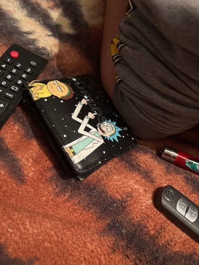The image depicts a scene on a reddish-brown patterned surface, possibly a rug or carpet. A gray sweatpants-clad leg is visible in the upper left. Scattered on the surface are a black remote control, a black Rick and Morty wallet, a silver and red lighter, and a black car key. No people are identified or discernible. The location is unknown as the image lacks indicators of Dobrovnitsa, Bulgaria.