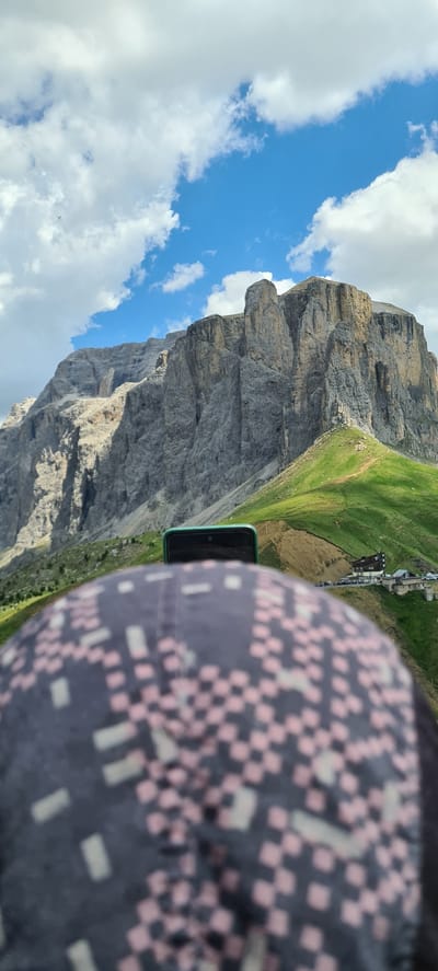This media file is a photograph taken from a high vantage point, looking down onto a dramatic mountain landscape. The primary subjects are the sheer, rocky face of a large mountain, a lush green grassy slope dotted with some buildings, and a segment of a person's head and shoulder, wearing a dark cap with a pink and cream checkered pattern. A smartphone with a teal-colored case is visible, held in front of the cap.
The scene appears to be set in the Italian Alps, specifically Sëlva - Wolkenstein - Selva di Val Gardena, as indicated by the context. The time of day is daytime, with a bright blue sky filled with fluffy white clouds. The weather seems pleasant and sunny.
The activity taking place is implied to be a scenic view or possibly a selfie being taken from a high altitude, perhaps on a hike, cable car, or a viewpoint. The person's head and shoulder being the closest element in the frame suggest they are the one taking the picture or are the subject of it. The detailed mountain face, the green slopes, and the buildings suggest a resort or a village nestled within the mountains. The perspective looking down the steep slope adds a sense of height and scale to the location. There is no visible text in the image.