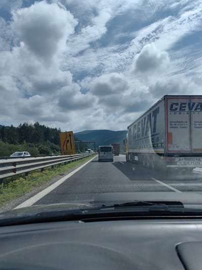 This media file depicts a scene on a highway under a cloudy sky, suggesting a daytime setting. The camera appears to be positioned inside a vehicle, looking forward through the windshield.
The main subjects are vehicles on the road. Prominently featured is a large white truck with "CEVA LOGISTICS" written on its side in large blue letters. Other vehicles visible include a white van and a red truck in the distance, as well as a silver car on the left side of the highway.
The highway has multiple lanes and is bordered by a metal guardrail and vegetation. A yellow traffic sign with arrows indicating lane changes and "500m" is visible on the right side of the road. The truck also displays various text, including website addresses, phone numbers, and location names like "ANKARA - TURKEY" and "TRABZON". License plates and other markings on the vehicles are also present.
The overall atmosphere is that of normal highway traffic. The cloudy sky suggests it might be overcast or that there's a mix of sun and clouds, but it is not raining. The presence of trees and hills in the background hints at a location outside of a dense urban area, possibly in a more rural or suburban highway setting. No people are clearly visible within the vehicles.