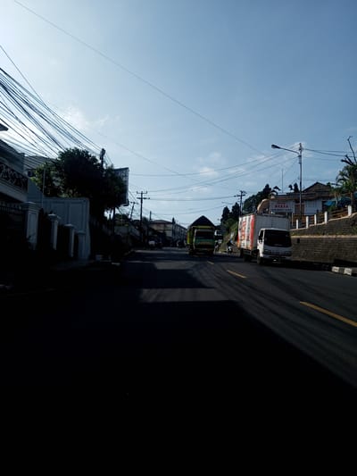 The image depicts a street scene in an urban environment, potentially Nhot Chang, Laos. The roadway is shadowed in the foreground, transitioning to sunlit areas further down the street. A yellow centerline marks the asphalt road. On the right side of the road, a white truck is moving, and on the left side, a green truck is moving. Overhead, there is an intricate network of power lines and utility poles. Buildings line both sides of the road, with a sign visible on the right building. A person driving a motorcycle also is present. The sky above is clear and blue, indicating a bright day.