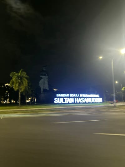 The image shows a night scene at the Sultan Hasanuddin International Airport in Baji Mangngai, Indonesia. A large, illuminated sign with the text "BANDAR UDARA INTERNASIONAL SULTAN HASANUDDIN" is prominently displayed. Behind the sign, a statue of a figure in traditional attire stands on a pedestal. Palm trees are visible to the left of the statue. The lighting in the image suggests it was taken at night, with streetlights and the illuminated sign providing illumination. The road in the foreground is visible, with motion blur indicating movement, possibly from a vehicle. The sky is dark with some clouds.