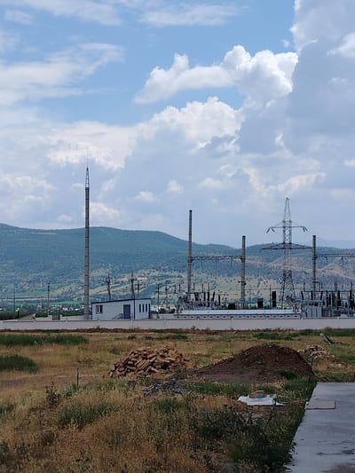 The image depicts an outdoor scene featuring a power substation situated in Gazalkent, Uzbekistan. In the foreground, dry grass and a pile of bricks are visible. The middle ground is dominated by the power substation, which includes a white building with a dark roof, multiple tall metal poles, electrical equipment, and power lines. A tall, thin tower with a pointed top stands to the left of the substation. In the background, green, rolling hills rise against a sky filled with white, fluffy clouds. The overall scene conveys an industrial landscape with natural elements. There are no people visible in the image.