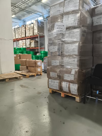 The image depicts a warehouse interior in Barysaw, Belarus. The scene is dominated by stacked boxes, predominantly cardboard, many of which are wrapped in clear plastic film. These boxes are arranged on pallets and industrial shelving units. In the foreground, a few empty wooden pallets are visible on the concrete floor, which shows signs of wear, including cracks and water stains. To the right, a black office chair is positioned near a stack of boxes. The background reveals more shelving units loaded with various goods, including green plastic containers. Overhead, a network of pipes and lighting fixtures are present. The overall impression is of a functional, albeit somewhat cluttered, storage facility.