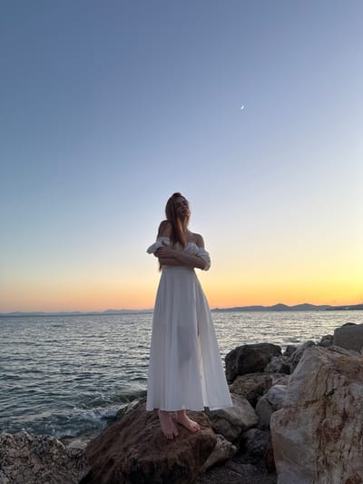 A woman stands on a rocky shoreline in Paleo Faliro, Greece, at dusk. She is wearing a flowing white dress and has her arms crossed in front of her. The sea is to her left, and the sky is a gradient of blue and orange, with a crescent moon visible. The rocks are varied in color and texture, with some appearing to be covered in lichen. The overall impression is one of serene beauty and contemplation.