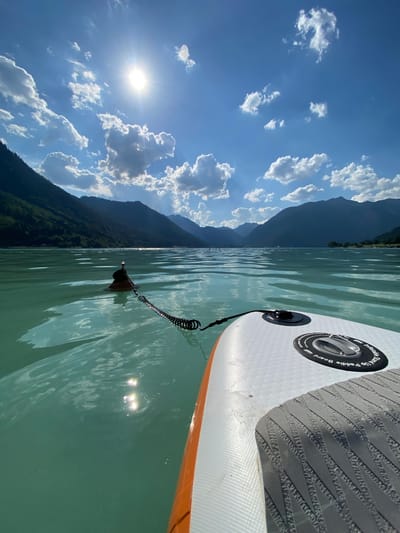 An eye-level, wide-angle, outdoor, daytime shot captures the view from a stand-up paddleboard on a body of water, likely a lake or fjord. The water is a striking turquoise hue, reflecting the bright sun and scattered white clouds in the vast blue sky. Majestic, tree-covered mountains form a dramatic backdrop, receding into the distance. In the foreground, the edge of the white and orange paddleboard is visible, along with its textured grip. A coiled leash extends from the board to a buoy floating in the water. The scene appears serene and tranquil, with the sun's rays creating shimmering patterns on the water's surface. The setting suggests a recreational activity in a natural, mountainous landscape.