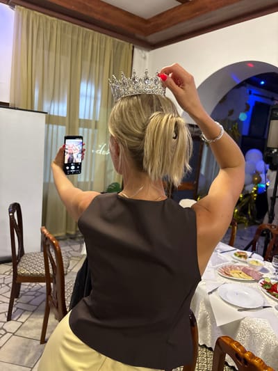 A woman with blonde hair is holding a tiara to her head while taking a selfie with her phone. She is wearing a dark top and a light-colored skirt. In the background, there are balloons and a table with food, suggesting a celebration or party. The lighting is dim, but there is some blue and red lighting from what appears to be decorative lights. The scene looks like an indoor event.