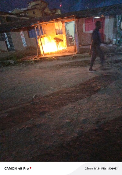 The image captures a nighttime street scene in Kitengela ward, Kenya, featuring a rudimentary open-fronted workshop alongside an unpaved road. In the workshop, an individual is actively engaged in a process generating intense orange sparks and light, consistent with metalworking (welding or grinding). This person is bent over an unseen object, silhouetted by the brilliant light. The workshop structure itself is constructed from corrugated metal sheets and wooden supports, with partially visible signage on its left exterior indicating "HARDWARE" and "DUKAVOO." To the right of the workshop stands another building, likely masonry, with a visible reddish-pink door or window. In the right midground, a second individual is captured walking from right to left across the unpaved, dusty ground, appearing blurred due to motion, and casting a prominent shadow. In the distant background, several multi-story buildings are silhouetted against the dark sky, with a faint red light visible at the top-left horizon. The overall environment is dimly lit by artificial light sources.
