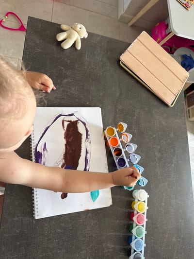 An overhead shot captures a young child's arm and shoulder as they paint on a spiral-bound notepad resting on a dark, textured countertop. The child is holding a paintbrush with their right hand, poised over a palette of watercolor paints in various shades of orange, yellow, blue, and purple. Beside the notepad, a small, cream-colored plush bear toy lies on the countertop. To the upper left, a bright pink toy stethoscope is visible. In the background, a tablet with a beige cover is placed on the countertop, and further back, a white table with pink and blue toy storage bins is partially visible. The scene appears to be an indoor setting, possibly a home or classroom in Верхнерусское, Russia, where the child is engaged in a creative activity.
