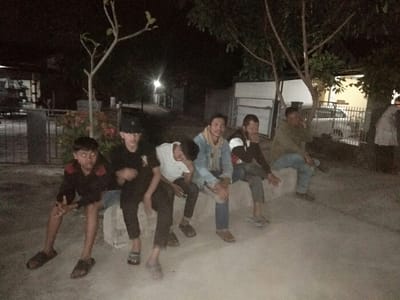 The image displays six males seated outdoors at night in what appears to be a residential street or communal area in Baleendah, Indonesia. They are positioned along a low, concrete wall or bench. The scene is dimly lit, indicating it is nighttime, with ambient artificial light sources providing illumination.

From left to right, the subjects include a young boy in a black shirt with red accents and sandals, notable for red-eye from the camera flash. Beside him is a male wearing a black cap, black t-shirt, black pants, and sandals. The third individual wears a white t-shirt, light blue jeans, and sandals, leaning forward with his head down. Next is a man in a light blue denim jacket over a checkered shirt, dark pants, and sandals. The fifth subject, with a beard, is dressed in a dark jacket and dark pants, looking away from the camera. The rightmost individual wears a light brown or olive jacket, blue jeans, and dark shoes, also looking away.

In the background, trees with varying foliage density are visible, along with shrubs featuring red flowers. Beyond these, elements of a residential street can be seen, including houses, fences, and a white car parked behind a gate. Faint lights in the distance suggest streetlights or lights from buildings. The group appears to be casually gathered, sitting together, possibly socializing or resting. No specific event or intense activity is evident. No text is visible in the image. The overall impression is a relaxed, informal gathering during the evening hours in a local neighborhood setting.