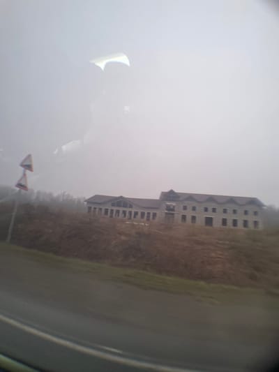 The image shows a tall, rectangular, multi-story building with many windows, likely under construction, on the edge of a grassy field, taken through a car window. A road sign is visible in the foreground. The weather appears foggy or overcast. The location is potentially in or near Bolgury, Russia.