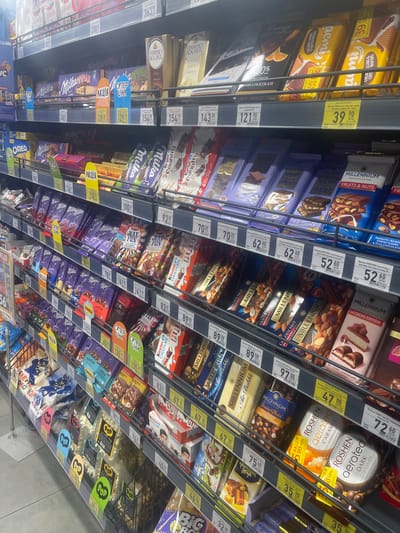 The image shows a shelf in a supermarket located in Horzhely, Ukraine. The shelf is filled with various chocolate bars and snacks. Brands visible include Milka, Kinder, Millennium, Roshen, and Oreo. There are also signs with prices and promotional tags like "Акція" (Promotion). The products are neatly arranged on multiple tiers of shelving. The lighting suggests an indoor commercial environment.