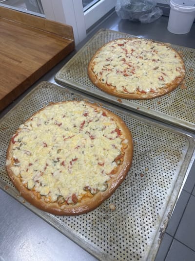 Two uncooked pizzas are displayed on perforated metal baking sheets. The pizzas are topped with melted cheese, tomato slices, and possibly other vegetables or meat. The baking sheets are placed on a stainless steel countertop, with a wooden cutting board to the left and a white plastic container and a plastic-wrapped item in the background. The scene appears to be in a commercial kitchen, potentially a pizzeria, in the city of Severodvinsk, Russia.