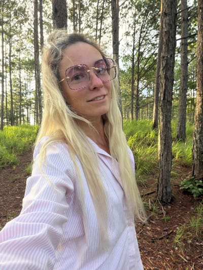 A blonde woman wearing large pink-tinted glasses and a pink and white striped button-down shirt is taking a selfie in a forest. Her hair is long and appears windswept. The background shows several tall, slender trees with rough bark, and lush green grass and pine needles on the ground. The lighting suggests it is daytime, possibly late afternoon due to the warm glow. The location is the city of Xuan Truong - Da Lat, Vietnam.