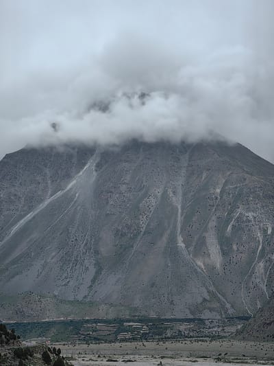 The image depicts a majestic mountain shrouded in heavy, grey clouds. The mountain's face is rugged and textured, with visible rocky outcrops and a stark, greyish-blue hue. Thin, lighter-colored streaks, likely representing watercourses or rock formations, are etched across its surface. At the base of the mountain, a valley unfolds, featuring a scattering of sparse vegetation, including small, dark-green trees and shrubs. A road curves along the left side of the valley, with what appears to be a red building and several other structures visible further in. The sky is overcast, creating a muted and somewhat somber atmosphere. The scene is likely located in or around Jispa, India, a region known for its dramatic mountainous landscapes.