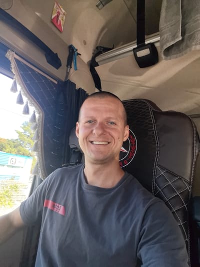 A man with a shaved head and a smile sits in the driver's seat of a truck. He is wearing a dark grey t-shirt with a red and black logo on the left chest. The truck's interior is visible behind him, with a black leather seat featuring white stitching and a circular red and white logo. There is a blue hose hanging from the ceiling on the left, and a grey towel is draped on the right. A picture of a couple in wedding attire is affixed to the ceiling. Through the window, trees and a light blue building with white lettering can be seen, suggesting an outdoor location in Saint-Herblain, France.