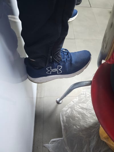 A person wearing blue Under Armour sneakers and dark blue pants is seated on a red chair with metal legs. The chair is positioned on a white tiled floor. The background includes a partially visible second shoe, what appears to be a plastic bag or wrap, and sections of grey tiled flooring. There is no verifiable information about Holywell, United Kingdom in the image.