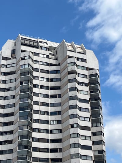 The image shows the exterior of a tall, modern apartment building in the city of Créteil, France. The building features a facade of white tiled panels and beige bands that create a staggered, geometric pattern. Numerous windows and balconies are visible on each level, indicating a residential structure. The building is set against a bright blue sky with scattered white clouds. The perspective is from below, looking up at the building. The scene is devoid of people or any discernible activity.