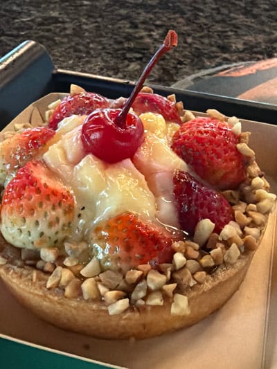 A close-up shot captures a single, circular pastry topped with sliced strawberries, a whole maraschino cherry on a stem, and chopped nuts. The pastry has a light brown crust and is filled with a creamy white substance that partially covers the fruit. The strawberries are a vibrant red, some sliced and others whole, with visible seeds. The cherry is a deep red, glossy, and has a green stem extending upwards. The chopped nuts are scattered around the fruit and the edges of the pastry. The pastry is presented in a light brown cardboard container. The background is blurred, showing a dark, textured surface on the left and a lighter, textured surface on the right, suggesting an indoor setting with some depth of field. The lighting is soft, highlighting the glossy surfaces of the fruit and the creamy filling. The overall impression is of a sweet, appealing dessert. No visible text is present.