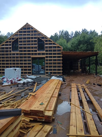 This image depicts an unfinished house under construction in a rural setting, likely in the Плешчаніцкі сельскі Савет, Belarus. The house's frame is exposed, showing wooden studs and black building wrap. Two windows and a large glass sliding door are visible on the front of the structure. A covered porch or extension is attached to the side of the house, with stairs leading up to it.
The ground around the house is a mixture of sand, gravel, and mud, with several puddles indicating recent rain. Construction materials are scattered throughout the scene, including stacks of lumber, bags of what appear to be insulation or building materials, and smaller piles of bricks or pavers. The lumber is predominantly light brown and appears to be unfinished.
The sky is overcast with thick, gray clouds, suggesting a cloudy or rainy day. The time of day is not definitively clear but the lighting suggests it is daytime, possibly late afternoon or early evening. Dense green trees surround the construction site, indicating a location in or near a forest.
There is no visible human activity in the image, making it a still shot of the construction process. Some bags of materials have visible text, though it is difficult to read clearly. One large white-wrapped object has some text that appears to be in a Cyrillic alphabet. The overall impression is one of ongoing construction in a natural, somewhat damp environment.