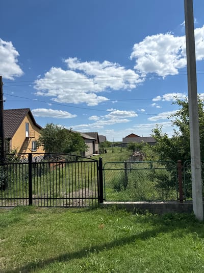 The image depicts a rural landscape under a bright, partly cloudy sky. In the foreground, a black wrought-iron fence stands partially open, revealing a grassy yard beyond. To the left, a yellow house with a dark brown roof and an external staircase is visible. Behind the fence, various structures are scattered across the property, including what appears to be a brick building and several other houses with tiled roofs in the distance. Lush green trees and vegetation are prevalent throughout the scene. A concrete utility pole stands prominently on the right side of the image, with wires running across the sky. The overall impression is that of a serene countryside setting, likely in the village of Dovhe, Ukraine, given the architectural style and the presence of utility poles and fencing common in such areas. No people or animals are present in the image.