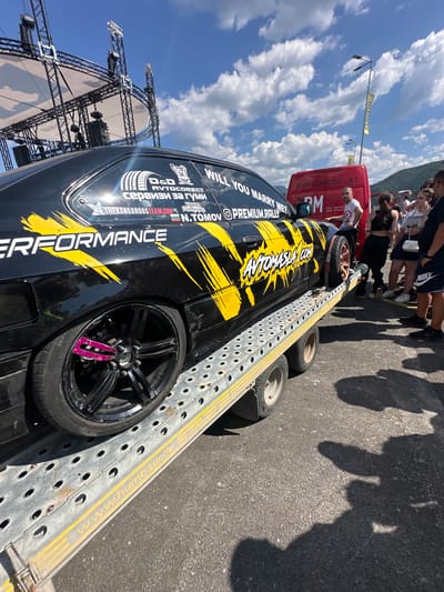 In Dospey, Bulgaria, a black car with yellow decals is positioned on a car trailer. The car displays various stickers, including "PERFORMANCE," "AVTOMASLA.COM," and "WILL YOU MARRY ME?". A red van with the website "www.pmcnc.at" visible on its side is parked nearby. Several people are gathered around the car and van, some looking at the car, while others are engaged in conversation. The scene appears to be outdoors, possibly at an event, with a stage structure and lighting rigs visible in the background under a partly cloudy sky. The trailer has yellow stripes and text that reads "www.humbaur.de."