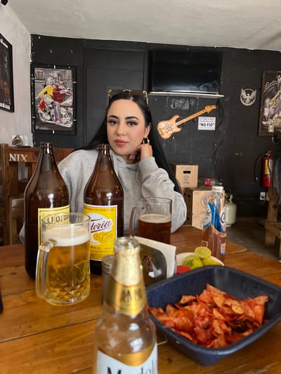 A woman poses at a table with several open bottles of beer and two full mugs. There's a blue bowl of chips nearby, along with limes, a small box and a few other bottles. The background features a dark wall adorned with art and a wooden guitar, creating a bar-like atmosphere.