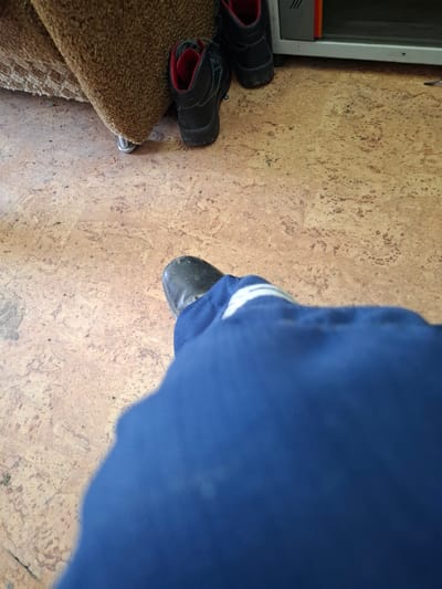The image depicts an indoor scene with a focus on footwear and a person's legs clad in blue trousers. In the background, a piece of furniture with a textured brown upholstery is visible, along with two pairs of black work boots. One pair of boots, positioned near the furniture, has red lining and appears to be worn. Another pair is further to the right, near a metallic and orange object, possibly a piece of equipment. The floor has a light brown, speckled texture. In the foreground, a person's right leg and foot, wearing a black boot and blue trousers with white stripes, is prominently featured, suggesting the image was taken from the perspective of someone sitting or resting. The overall scene appears to be a functional or work-related environment. The location is identified as Dergachi, Russia.