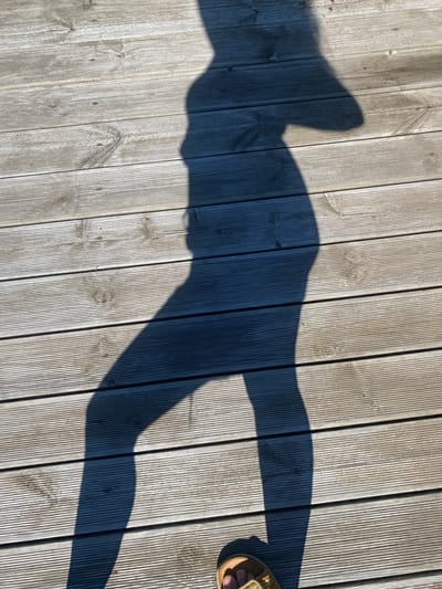 A shadow of a person wearing a wide-brimmed hat and sandals is cast across a wooden deck. The shadow is positioned in the center of the frame, with the person's legs extending towards the bottom of the image. The wooden planks of the deck are oriented horizontally and show signs of wear and weathering. The scene is outdoors, under bright sunlight, as indicated by the distinct shadow. The location is stated to be in the city of Bol'shoe Zaborod'e, Russia.