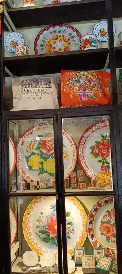 A multi-tiered wooden shelf is filled with colorful vintage items. Enamel plates with floral designs dominate the display, some behind glass doors. A beige tote bag with "ARLENE HOUSE" printed on it sits beside an orange pillow with a floral pattern. Small bottles and boxes of medicines are arranged neatly alongside the enamel plates behind the glass, adding to the nostalgic feel. There are also white stones and boxes.