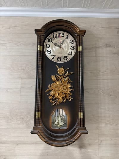 A grandfather clock is mounted on a light-colored wood-paneled wall. The clock has a dark brown wooden case with ornate gold detailing. The clock face is round with black Arabic numerals and a gold frame. A gold floral ornament is affixed to the front of the clock, below the face. A glass window at the bottom of the clock reveals a pendulum and chimes. The ceiling above the clock is white and textured. The environment appears to be indoors.