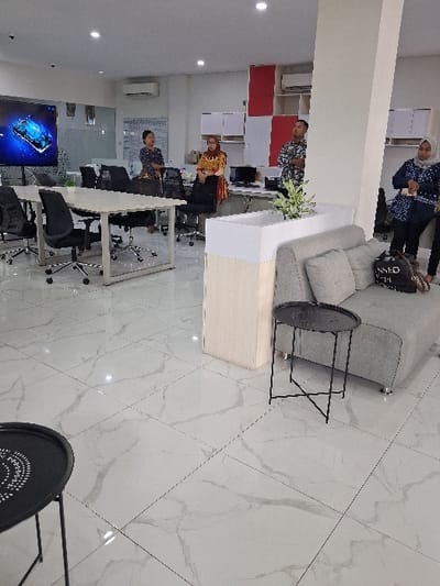 A brightly lit office features white marble flooring. A gray couch sits near a white and light-colored wood reception desk. Several people occupy the space. Two black side tables are placed around the couch. Desks and chairs are positioned in the background, with a large screen on the wall displaying an image. The layout suggests a modern and functional workspace.