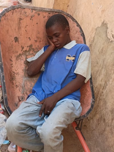 A young individual, identified as male, is depicted seated within the orange-red, heavily rusted metal tub of a wheelbarrow, which functions as a makeshift chair. The scene is located in Kawaji, Nigeria. The male, with dark skin and short black hair, is positioned with his eyes closed, head resting on his right hand, suggesting sleep or deep rest. He wears a short-sleeved blue polo shirt featuring light beige/off-white sleeves and collar, with a yellow "M" logo and "AMARSHA" text on the left chest. Light grey trousers are worn, and a silver-colored ring is visible on the ring finger of his left hand. The immediate background to the right consists of a light brown wall exhibiting wear and possible damage. The ground beneath the wheelbarrow shows scattered debris, likely plastic materials, on a dirt surface.