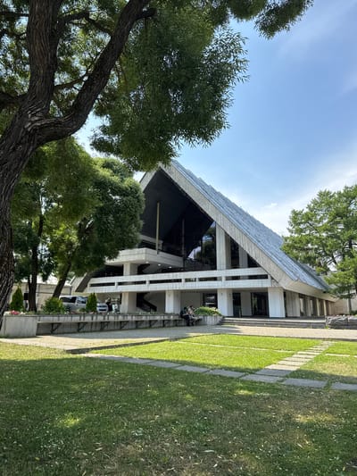 A modern, angular building with a large, sloping roof is the central focus of the image. The building appears to be made of concrete and glass, with multiple levels and large windows. It is surrounded by greenery, including large trees and a well-maintained lawn. Several people are visible, some sitting on benches and others standing near the entrance. The sky is clear and blue, suggesting a sunny day. The scene is quiet and serene, with no obvious signs of activity.