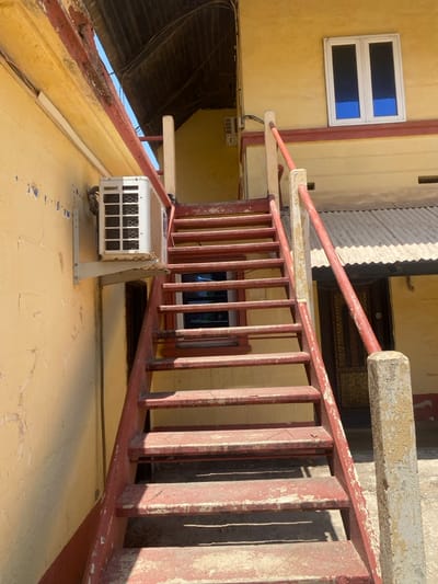 The scene, located in Tudun Nupawa, Nigeria, depicts an outdoor residential or commercial building facade under bright daylight. A prominent, weathered, rust-red painted metal staircase with concrete newel posts and metal handrails ascends centrally towards the upper right. The staircase exhibits significant paint peeling and surface degradation.
To the left of the staircase, a light yellow-painted wall, showing cracking and peeling paint, features a white outdoor air conditioning condenser unit mounted on a metal bracket at mid-height. Below the unit, a dark rectangular opening, possibly a doorway or recessed area, is visible. Above the staircase, the upper section of the building also presents a faded yellow facade, adorned with a white-framed, two-pane window containing dark blue tinted glass.
To the right of the staircase, partially visible beneath an overhang, is a section of a lower building with a grey corrugated metal roof. A dark entryway with a discernible patterned metal grate is situated beneath this roof structure. Above the upper left portion of the staircase, exposed wooden joists or the underside of a roof structure are visible. The ground surface at the base of the staircase is a light-colored, possibly concrete, patio area. No individuals or active events are present in the image. Shadows cast by the staircase elements indicate strong direct sunlight.