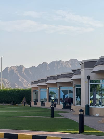 This image captures a tranquil outdoor scene at a resort or residential complex in Al Fiqait, United Arab Emirates, with arid mountains forming a dramatic backdrop.
In the foreground, a vibrant green, meticulously maintained lawn stretches across the lower right, bordered by a paved walkway. Along this path are black bollard lights with domed tops, providing illumination. A black and yellow striped safety barrier is visible on the far right edge. Closer to the building, a dark green waste bin is situated on the paved area.
On the right side of the frame, a modern multi-story building features light-colored walls and tiered dark brown roof tiles. It contains multiple units, each with large windows reflecting the surrounding environment, including palm trees and the sky. Some units have private patios or balconies, equipped with outdoor tables and chairs. Small golden plaques with unit numbers like "A-103," "B-201," and "B-202" are visible next to some entrances.
The central and left portions of the image are dominated by rugged, rocky mountains, characteristic of the region. These mountains appear somewhat hazy, suggesting distance or atmospheric conditions. The sky above is a soft, pale blue, with delicate, wispy white clouds scattered sparsely. A few tiny dark specks, possibly birds, can be seen against the bright sky.
No people are visible, contributing to the serene and peaceful ambiance. The soft lighting suggests either early morning or late afternoon. The weather appears clear and calm, with a gentle haze over the distant mountains.