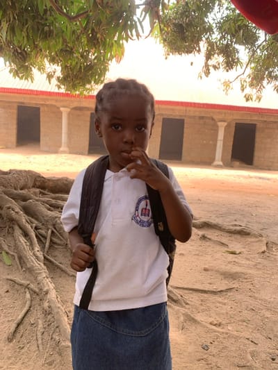 A young child, appearing to be female, stands in an outdoor environment in Garaku, Nigeria. The child has dark skin and hair styled in braids, looking directly at the viewer with a neutral expression. She is wearing a white short-sleeved polo shirt, a dark backpack worn over both shoulders, and a blue denim skirt. A visible emblem on the left chest of the polo shirt contains text including "KUDO MODERN" and "GARAKU," along with an icon depicting an open book and a torch. The child's right hand is raised, with fingers positioned near the mouth. In the foreground to the left, a dense cluster of exposed tree roots is visible on the earthen ground. Overhead, green leaves from a tree cast dappled light. In the background, a single-story building constructed of light-colored block or brick, featuring a red corrugated metal roof, is present. The building exhibits multiple dark, rectangular door or window openings and at least one white fluted column. The ground throughout the scene is composed of bare earth or sand.