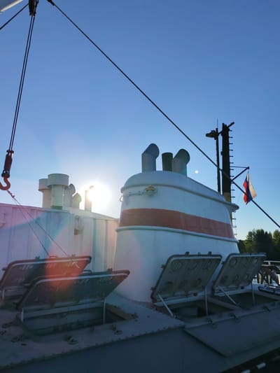 An upward-angled outdoor photograph depicts the deck and superstructure of a white vessel, likely a riverboat, located in Rodino, Russia, under a clear blue sky. The sun, positioned on the left side of the frame, generates significant glare and lens flare. A distinct, small white disc, consistent with the moon, is visible in the sky above the vessel's central structure, to the right of the sun's glare.
The vessel's primary superstructure is white with a prominent horizontal orange-red band and features multiple metal exhaust stacks. An adjacent white structure with additional vents is visible on the left. In the foreground on the deck, two rectangular metal hatches are open, propped upwards by struts, with a third closed hatch visible directly below them.
Dark rigging lines or cables traverse the sky diagonally; one line on the far left includes a heavy metal hook. On the right, a dark, vertical mast-like structure with ladder rungs extends upwards. A tattered Russian flag, displaying horizontal white, blue, and red stripes, flies from this mast. Distant green trees are visible along the horizon on the far right. No people are present.