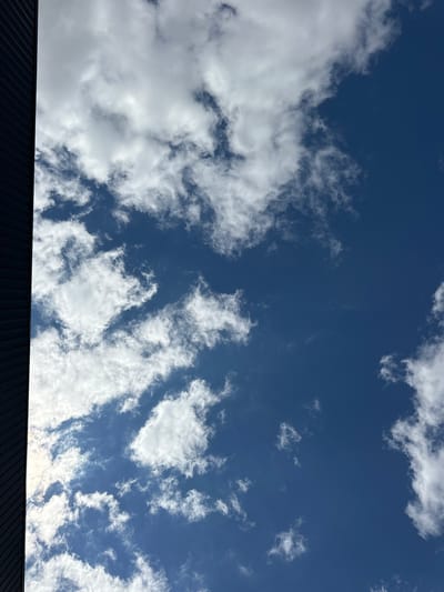 The image shows a clear blue sky with scattered white clouds. On the left side of the frame, a dark, ribbed structure, possibly a building facade, extends vertically from the bottom to the top of the image, obscuring a portion of the sky. The lighting suggests it is daytime, with sunlight illuminating the clouds from above. There are no people or specific objects clearly identifiable within the scene besides the sky and the partial view of the structure. The composition is a low-angle shot looking upwards. The image does not provide any contextual information that would suggest a specific location within the city of Saran, France.
